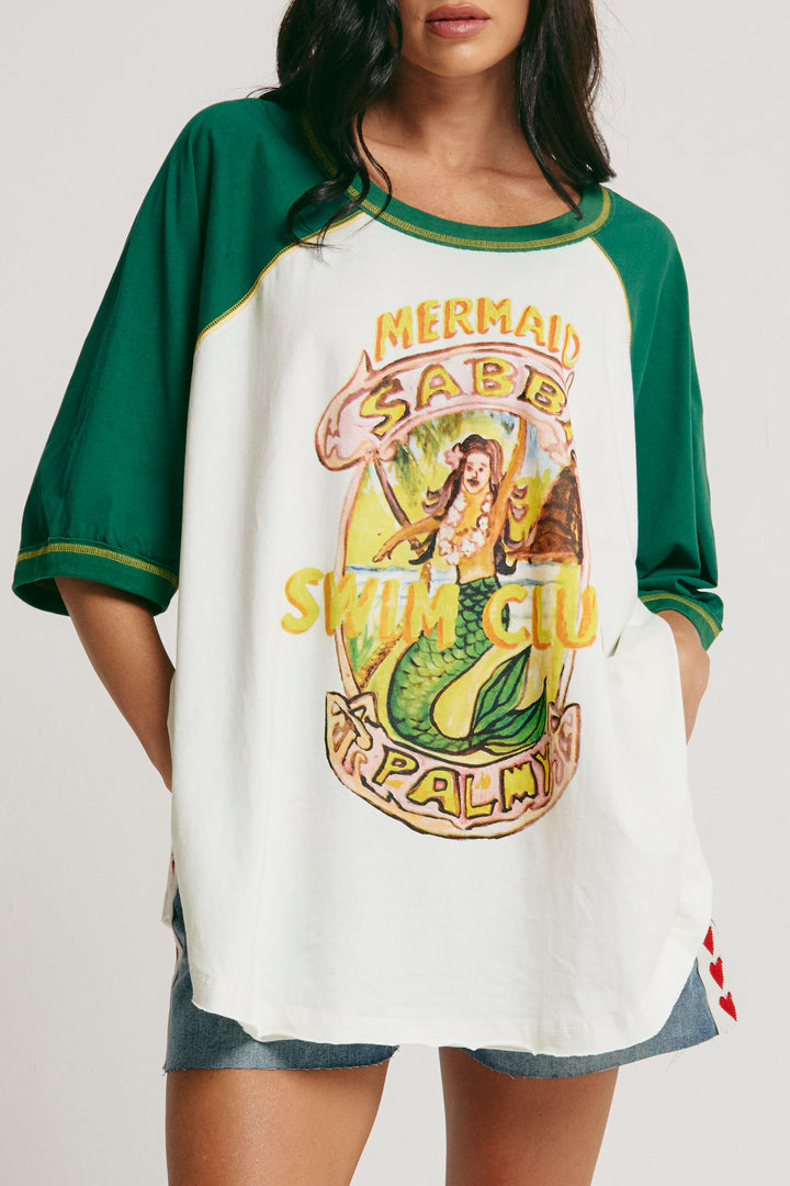 Person standing in photo studio wearing marshmallow and green Mermaid Swim Club Raglan Tee