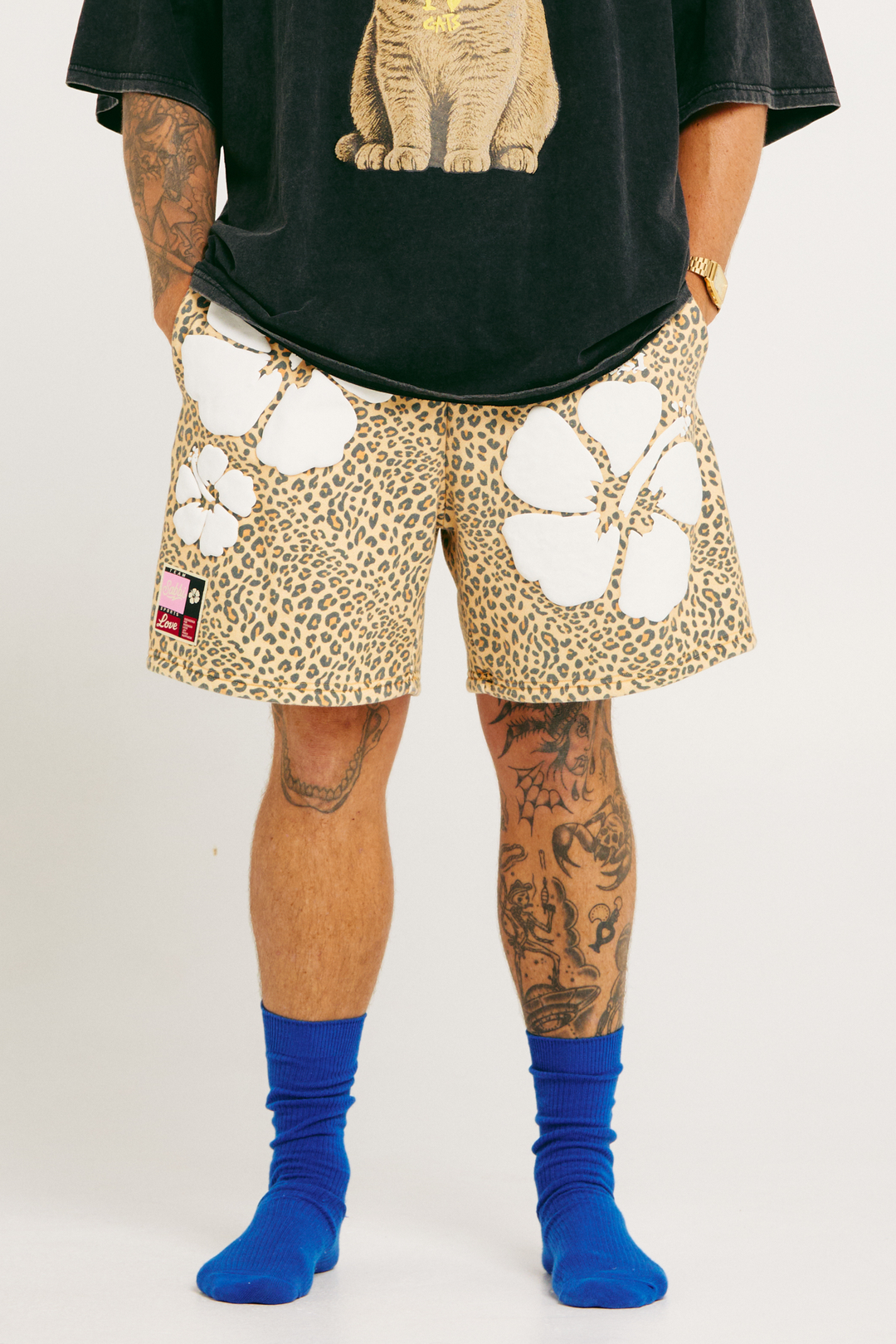 Person wearing leopard print shorts with white hibiscus patches, black shirt and blue socks on a plain background