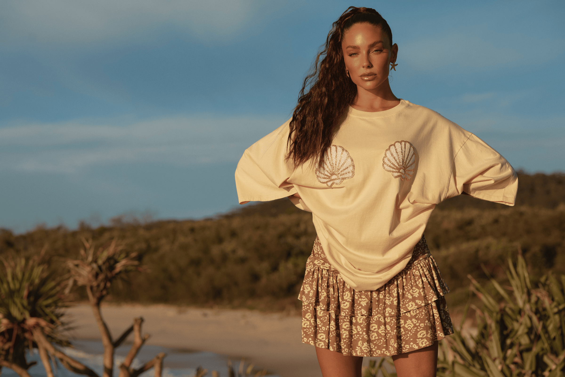 Woman wearing a beige t-shirt with a shell design and a floral skirt on a beach.