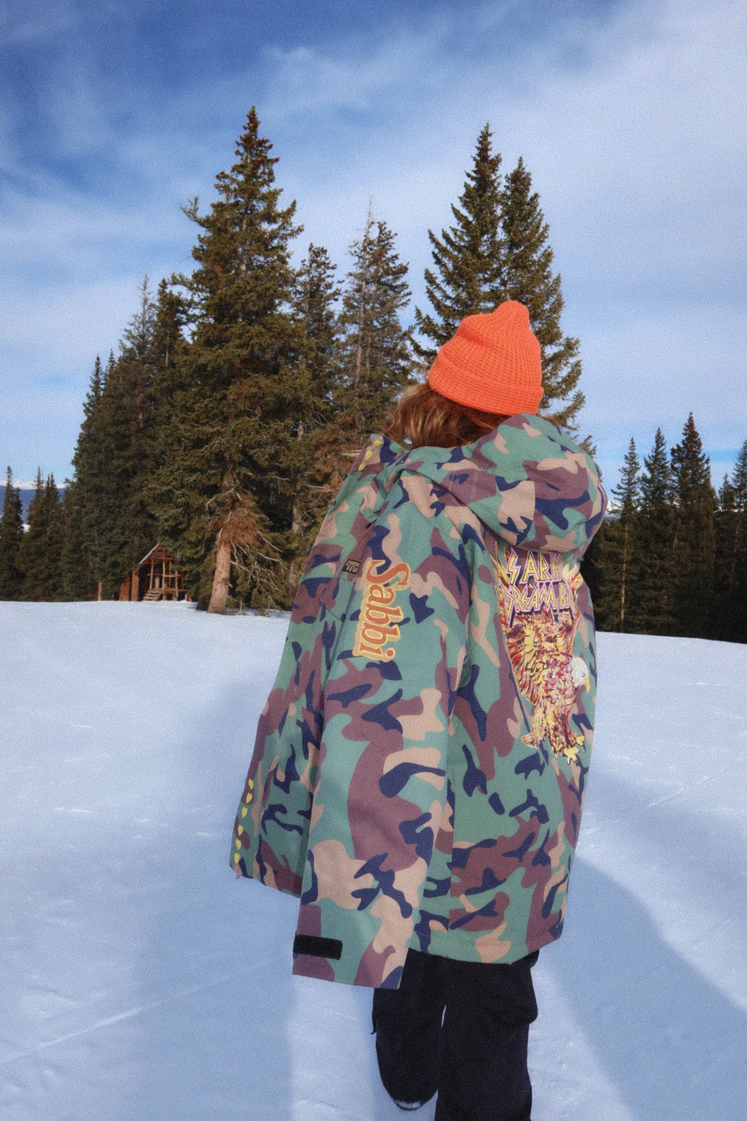 Person wearing a colorful jacket and orange beanie standing on snow with trees in the background