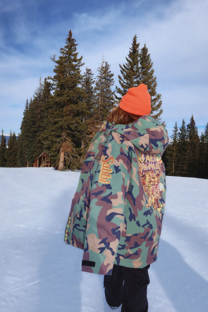 Person wearing a colorful jacket and orange beanie standing on snow with trees in the background