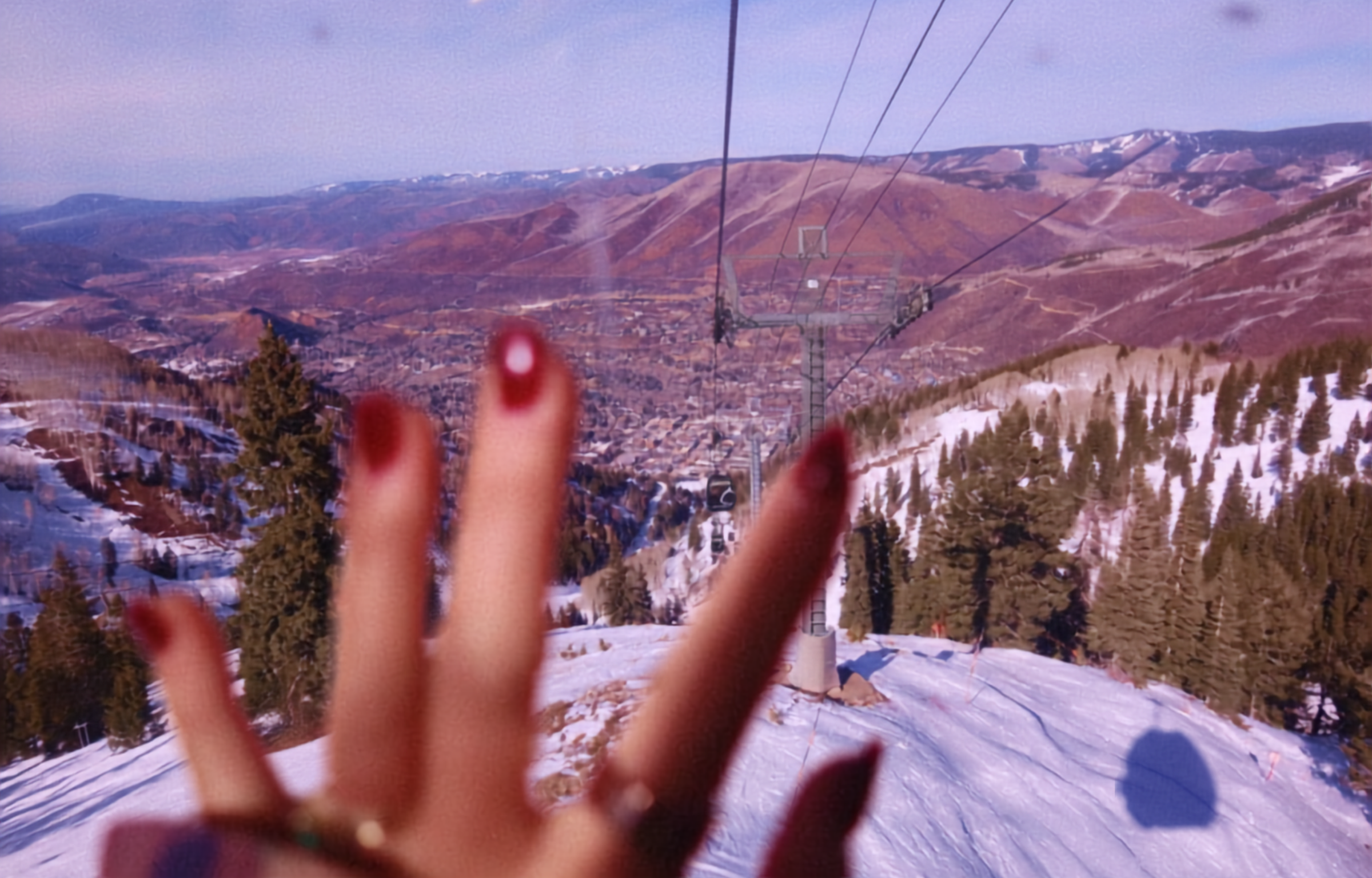 Hand with red nail polish blocking a scenic view of a ski resort and mountains.